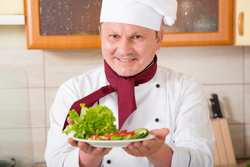 Mature male cook in the kitchen holding a plate of fresh vegetable salad. Prepares diet food. Lean dish.