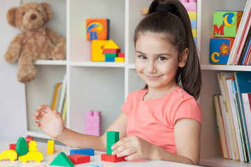 Back to school and happy time! Happy cute child girl sitting at a table with a white bookcase with colored books and toys behind. 