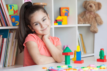 Back to school and happy time! Happy cute child girl sitting at a table with a white bookcase with colored books and toys behind. 