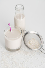rice milk in bottle and glass with straw near scattered rice and sieve on grey background