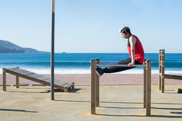 Young boy, practicing sports, calisthenics, with his headphones, outdoors, near the coast, with the blue sea in the background.