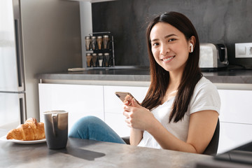 Portrait of adorable asian girl holding mobile phone while having breakfast in kitchen at home