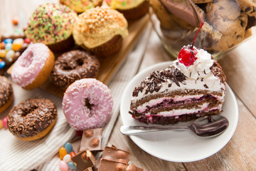 junk food, sweets and unhealthy eating concept - close up of glazed donuts, chocolate, drop candies and piece of cake on wooden table