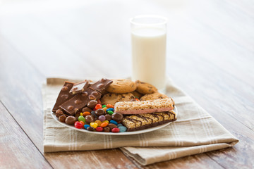 food, junk-food and unhealthy eating concept - close up of chocolate, oatmeal cookies, drop candies with muesli bars on plate and glass of milk