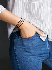 Cropped half-turn shot of woman's hand, wearing black lucky rope bracelet with 3 rows of thick string. The girl is wearing jeans and stripy shirt, putting hand into pocket, posing on dark background.