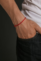 Cropped closeup shot of man's hand with tanned skin, wearing red lucky rope bracelet. The guy is wearing black jeans and white shirt, putting his hand into the pocket, posing on the dark background.