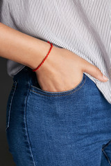 Cropped closeup shot of woman's hand, wearing red lucky rope bracelet with thick string. The girl is wearing jeans and stripy shirt, putting her hand into the pocket, posing on the dark background.
