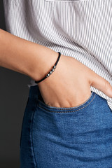 Cropped closeup shot of woman's hand, wearing black beaded lucky rope bracelet with thick string. The girl is wearing jeans and stripy shirt, putting hand into pocket, posing against dark background.