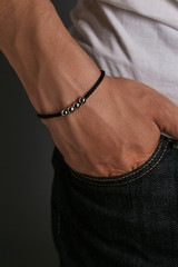Cropped closeup shot of man's hand with tanned skin, wearing black lucky rope bracelet with silver beads. The guy is wearing black jeans and white shirt, putting hand into pocket on dark background.