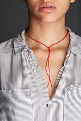 Cropped shot of woman's chest, wearing red cord necklace with thick string. The lady with tanned skin is wearing stripy shirt with top pockets, posing on dark background. Voguish women's accessory.