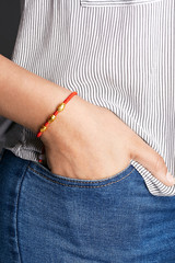 Cropped closeup shot of woman's hand, wearing red lucky rope bracelet, adorned with 3 golden beads. The girl is wearing jeans and stripy shirt, putting hand into pocket, posing on dark background.