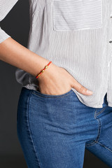 Cropped half-turn shot of woman's hand, wearing red lucky rope bracelet, adorned with 3 golden beads. The girl is wearing jeans and stripy shirt, putting hand into pocket, posing on dark background.