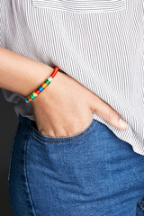 Cropped closeup shot of woman's hand, wearing red lucky rope bracelet with segmented winding. The lady is wearing jeans and stripy shirt, putting hand into pocket, posing against dark background.