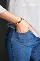 Cropped half-turn shot of woman's hand, wearing red lucky rope bracelet with segmented winding. The lady is wearing jeans and stripy shirt, putting hand into pocket, posing on the dark background.
