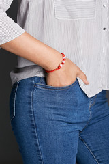 Cropped half-turn shot of lady's hand, wearing red lucky rope bracelet, adorned with 3 milky beads. The girl is wearing jeans and stripy shirt, putting hand into pocket, posing on dark background.