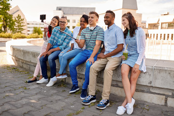 Fototapeta premium people, technology and friendship concept - group of happy international friends or students taking picture by smartpnone on selfie stick in city