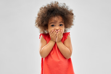 childhood, expression and emotion concept - confused little african american girl covering mouth by...