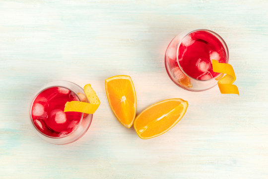 Two cocktail glasses with campari and orange twist garnishes, with ice cubes and orange wedges, shot from above on a white wooden background with copyspace