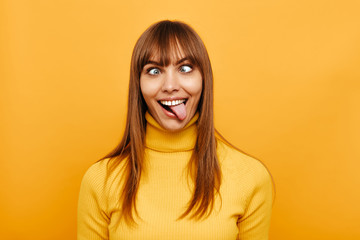 Woman portrait. Fun. Cheerful young woman is grimacing and showing her tongue at camera, on a yellow background - obrazy, fototapety, plakaty