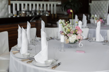 white napkins stand in white plates and bouquet of fresh flowers in the center of served table in the restaurant. clean white dishes layout on a white tablecloth