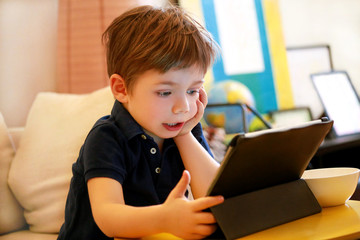 Child using tablet pc on bed at home. Cute boy on sofa is watching cartoon, playing games and learning from laptop. Education, fun, leisure, happiness, modern computer technology and communication.