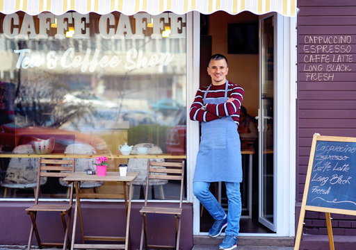 Portrait Of A Confident Young Man Standing In The Doorway Of A Coffee Shop. Tea And Coffee Shop Written On The Window. Traffic Reflection. His Welcoming Smile Makes A Great First Impression