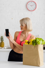 cheerful blonde woman in sportswear holding smartphone with blank screen near paper bag with groceries