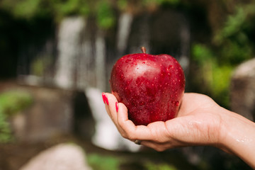 female hand holds a fresh tasty red apple on a background of a waterfall
