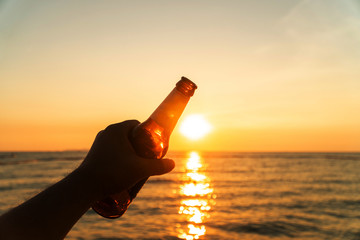 Hand of man is holding beer bottle and holds his hand up on the sky in evening with sunset....