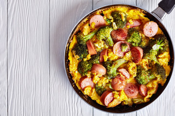delicious hot Omelette with broccoli and sausages in a skillet on a white wooden table, horizontal view from above, flat lay, close-up, copy space