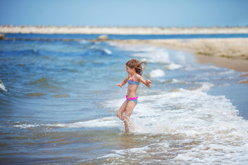 Fototapeta na wymiar Cute little girl runing along the seashore against a clear blue sky and rejoices in the rays of the summer sun