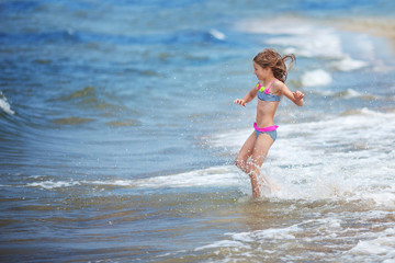 Cute little girl runing along the seashore against a clear blue sky and rejoices in the rays of the summer sun