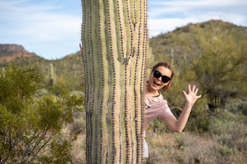Adult woman hides and waves while standing behind a large saguaro cactus in the Sonoran Desert - obrazy, fototapety, plakaty