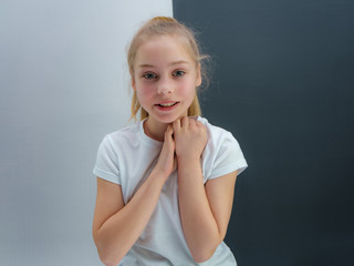 A concept portrait of a cute pretty blonde teen girl with long hair is sitting in a white T-shirt against a gray-white background and talking.