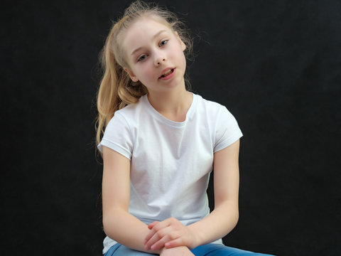 A concept portrait of a cute pretty blonde teen girl with long hair sitting in a white t-shirt against a black background in the studio and talking.