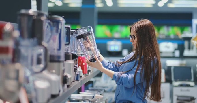 In the appliances store, a brunette woman in a shirt chooses a blender for shopping by viewing and holding the device in her hands.