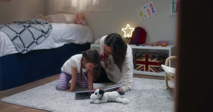 DOLLY Portrait of cute little girl reading a book together with her mother at home, evening shot, bedroom interior. 4K UHD RAW FOOTAGE