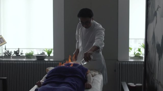 1080p video of an asian man pouring liquid on a fire lit on a towel over the lying person during a chinese fire massage