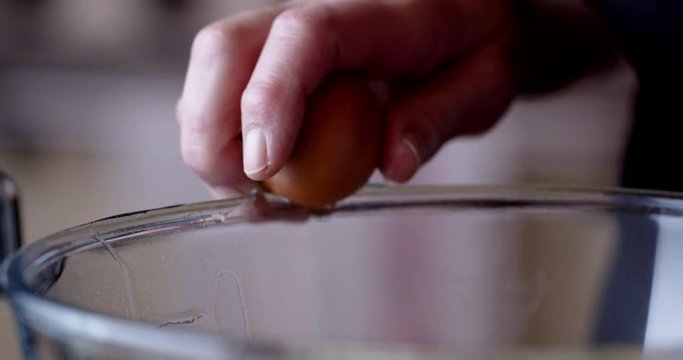 Slow motion and up close cracking of an egg on the side of a glass bowl. Egg is cracked and opened one-handed and falls into the bowl.