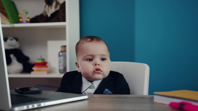 Adorable serious baby boss in business costume sitting on the chair at the table with laptop and smartphone in the office. Child�s portrait. Joke, having fun, work hard play hard, happy childhood