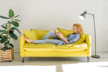 Woman lying on cozy yellow couch and reading book