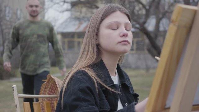 Pretty young woman painter sitting in front of wooden easel drawing a picture. Handsome man comes closer, covers eyes of the artist and hugs his girlfriend. Creativity, self-expression, love concept