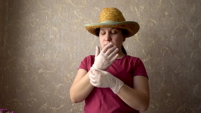 A woman puts hands on transparent gloves. On his head a straw hat. Preparation for work. Burgundy t-shirt. Gray Wallpaper on the wall. Home-room. 4K video.