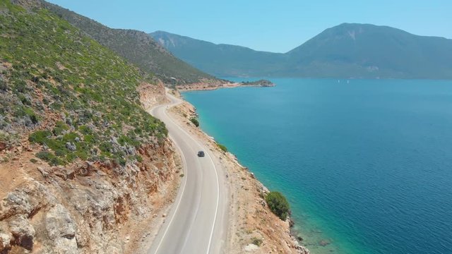 DRONE: Flying behind a tourist car exploring the remote island in the Mediterranean by driving down a scenic coastal road. Tourists cruising along the breathtaking shoreline of Lefkada in a grey car.