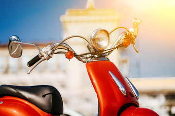 Closeup chrome detail and headlamp of orange retro vintage scooter under blue sky and sun in front of defocused tower