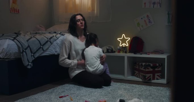 Portrait of young attractive mother cuddling and soothing her daughter at home, evening shot, bedroom interior. 4K UHD RAW FOOTAGE