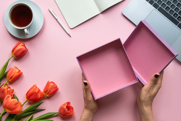 Female hands with open empty box on pink background. Background with coffee, laptop and flowers