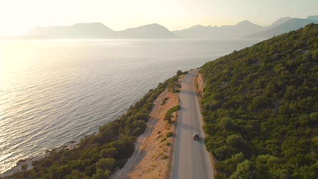 DRONE: Flying behind tourist car driving down empty road leading around the picturesque Greek island. Golden morning sun rays illuminate the Mediterranean nature and car cruising along the calm sea.