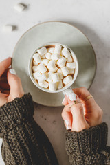 Girl's hands hold a cup of hot chocolate with marshmallow over textured white background. Top view, flat lay.