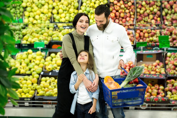 Portrait of a cheerful family standing with a full cart in the supermarket in the vegetable Department on the background of the counter with apples.